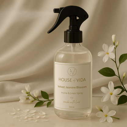 A clear glass bottle with a black spray nozzle labeled 'HOUSE OF VIDA Velvet Jasmine Blossom Home & Linen Spray 500ml', surrounded by white jasmine flowers and scattered petals.